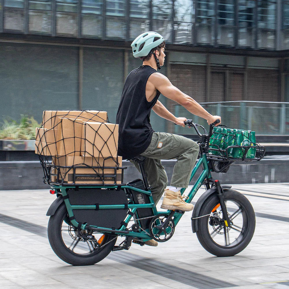 Person riding a compact cargo e-bike through a city plaza, carrying boxed goods and drinks for urban delivery.