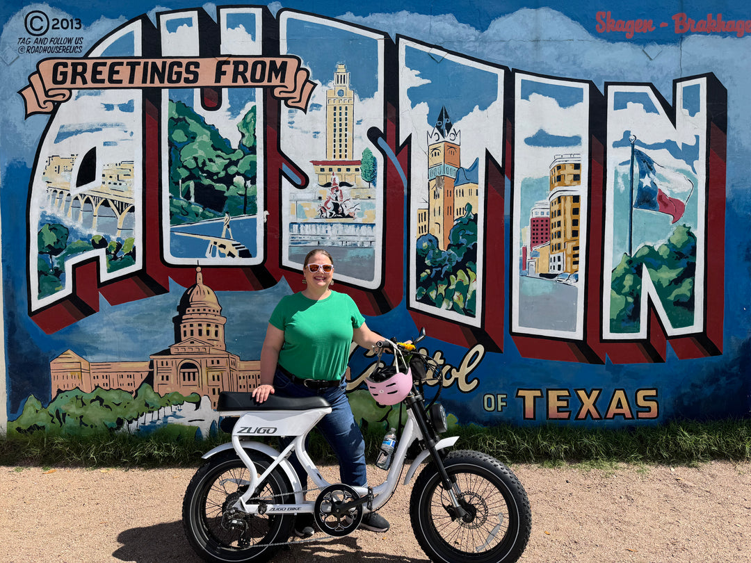 Rovo founder smiling in front of the iconic ‘Greetings from Austin’ mural with a white Zugo electric bike and pink helmet.